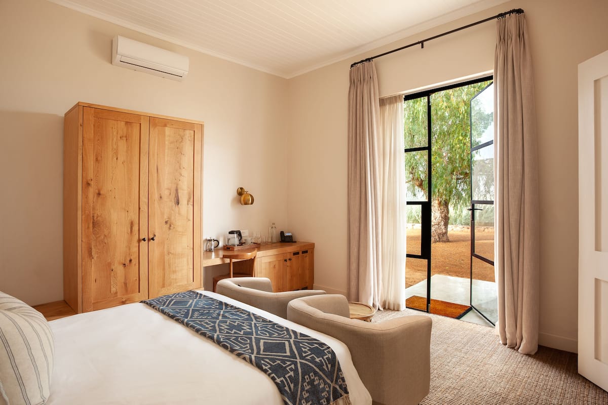 A bedroom with a bed, a wooden wardrobe, a desk with a chair, and a view of the outdoors through a glass door.