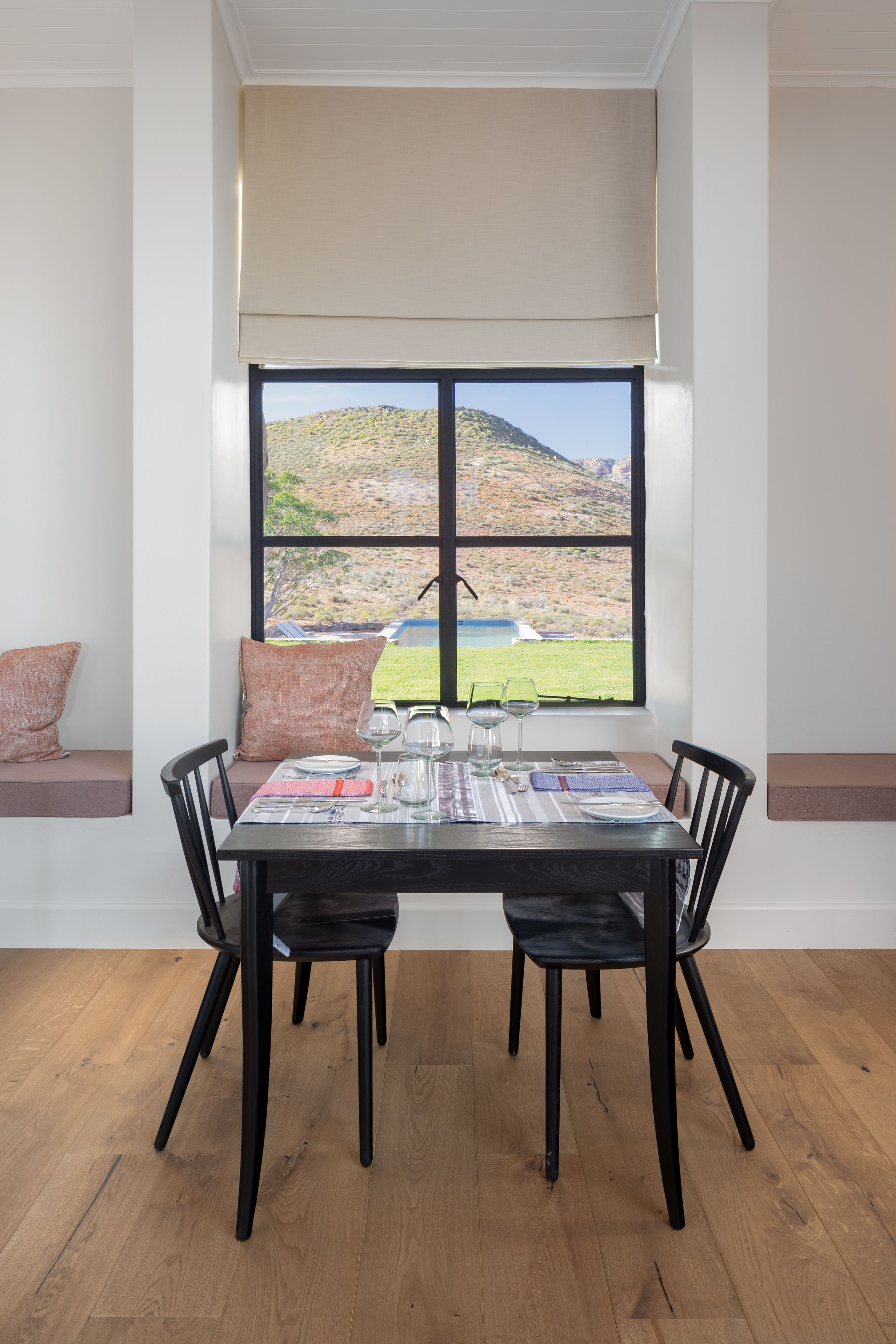 Un coin repas avec une table dressée pour deux personnes, une fenêtre donnant sur un paysage vallonné et un banc agrémenté de coussins.