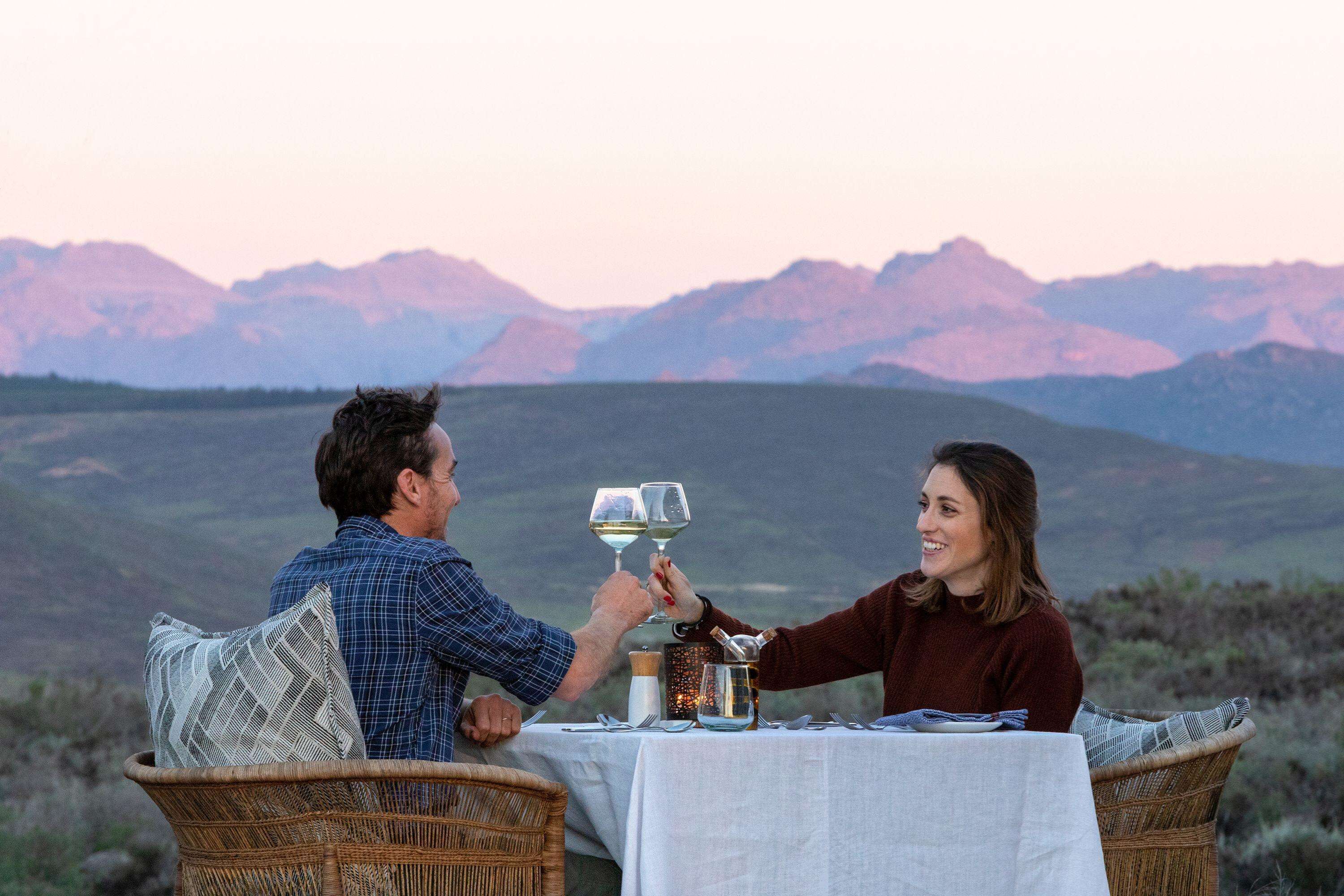 Um casal brindando com taças de vinho à mesa, com vista para as montanhas ao fundo.
