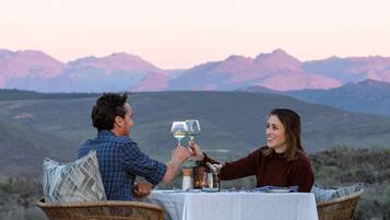 Um casal brindando com taças de vinho à mesa, com vista para as montanhas ao fundo.