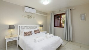Una cama bien hecha con sábanas blancas y almohadas decorativas, una mesita de noche con lámpara, y una ventana con cortinas.