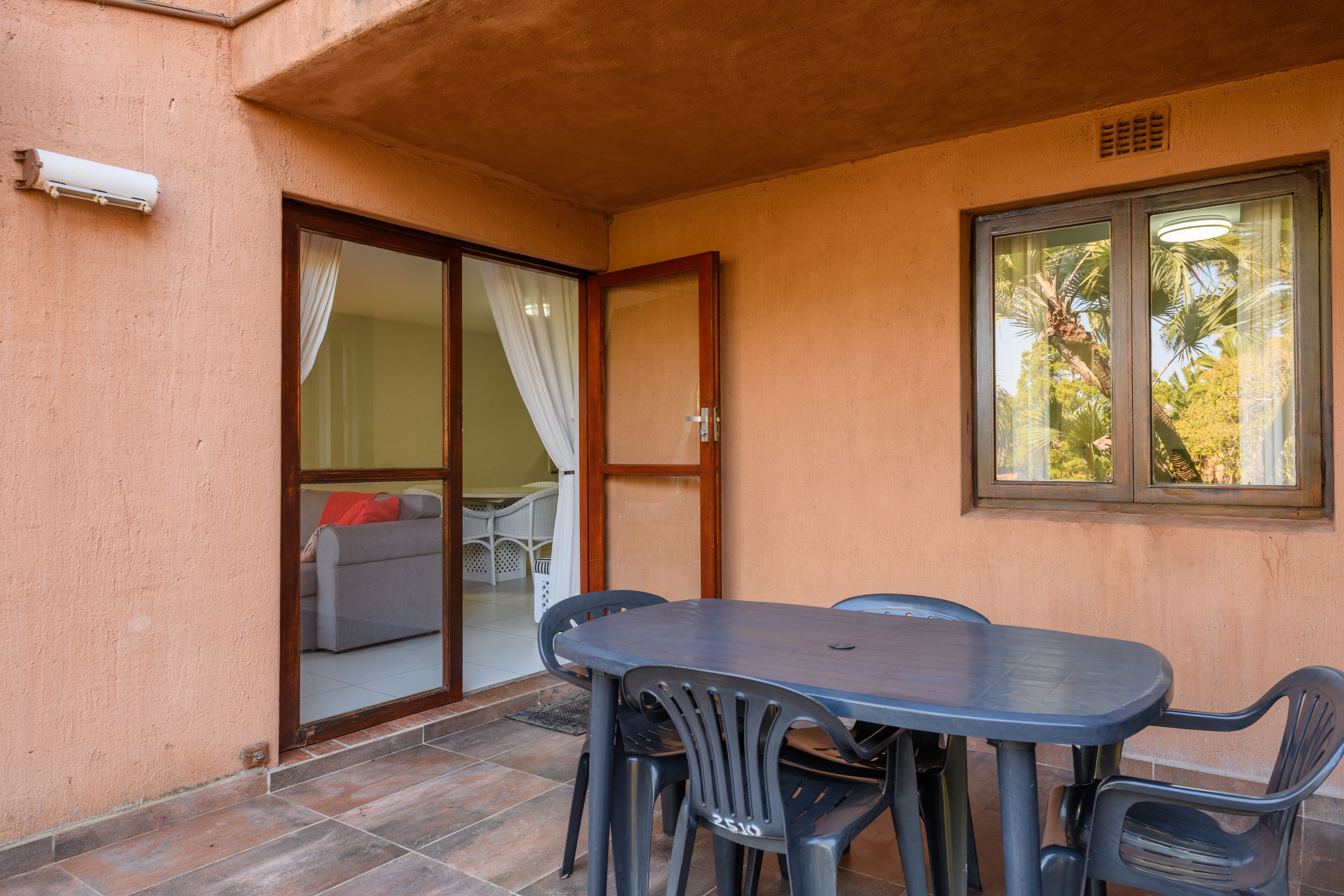 Un patio con una mesa y sillas, un patio con puerta corrediza que da a una habitación con sofá y cama, y una ventana con vista a palmeras.