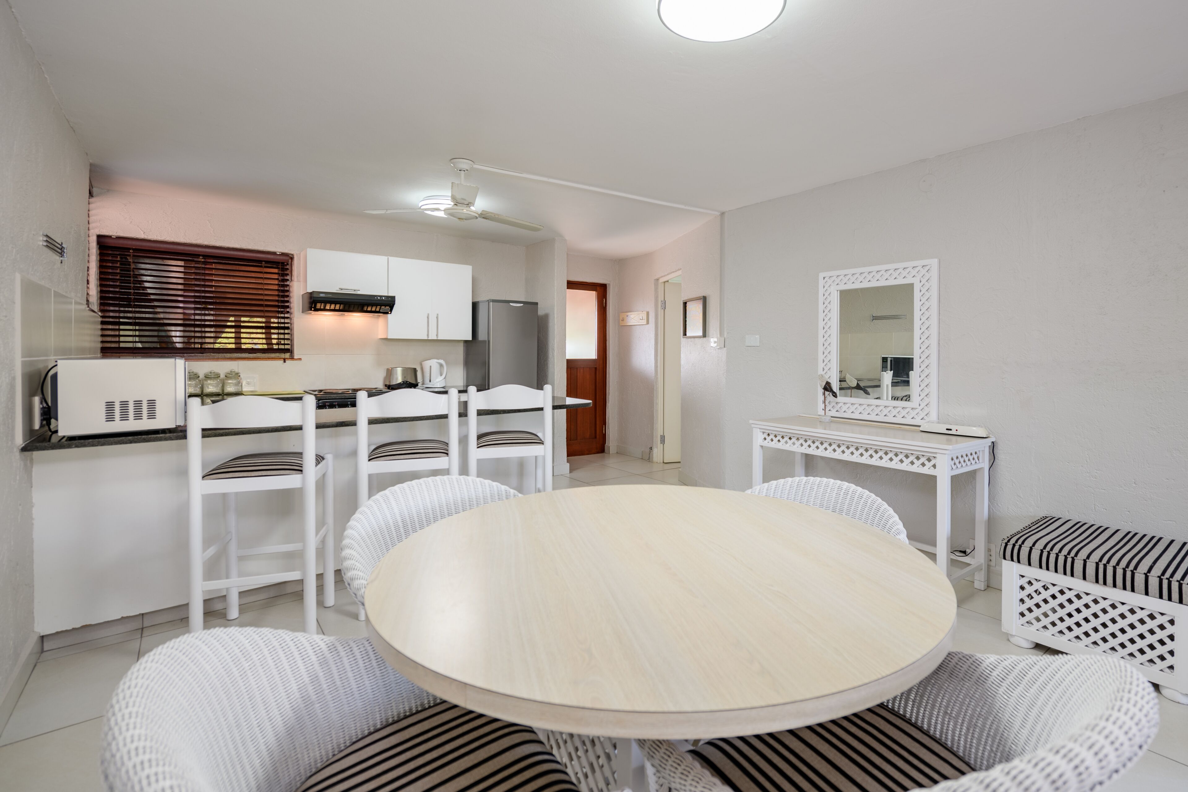 A compact kitchen with white cabinetry, a refrigerator, and a microwave. There is a dining area with a round table and white wicker chairs.