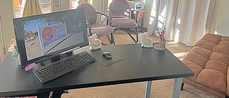 A computer desk with a monitor, keyboard, and mouse. There are two chairs, a small table with a vase and a Buddha statue, and a couch. A TV is mounted on the wall above the desk.
