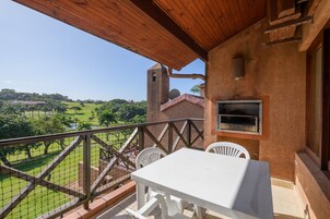 Un balcón con una mesa y sillas blancas, un muro de ladrillo y vistas a un paisaje verde con una torre.