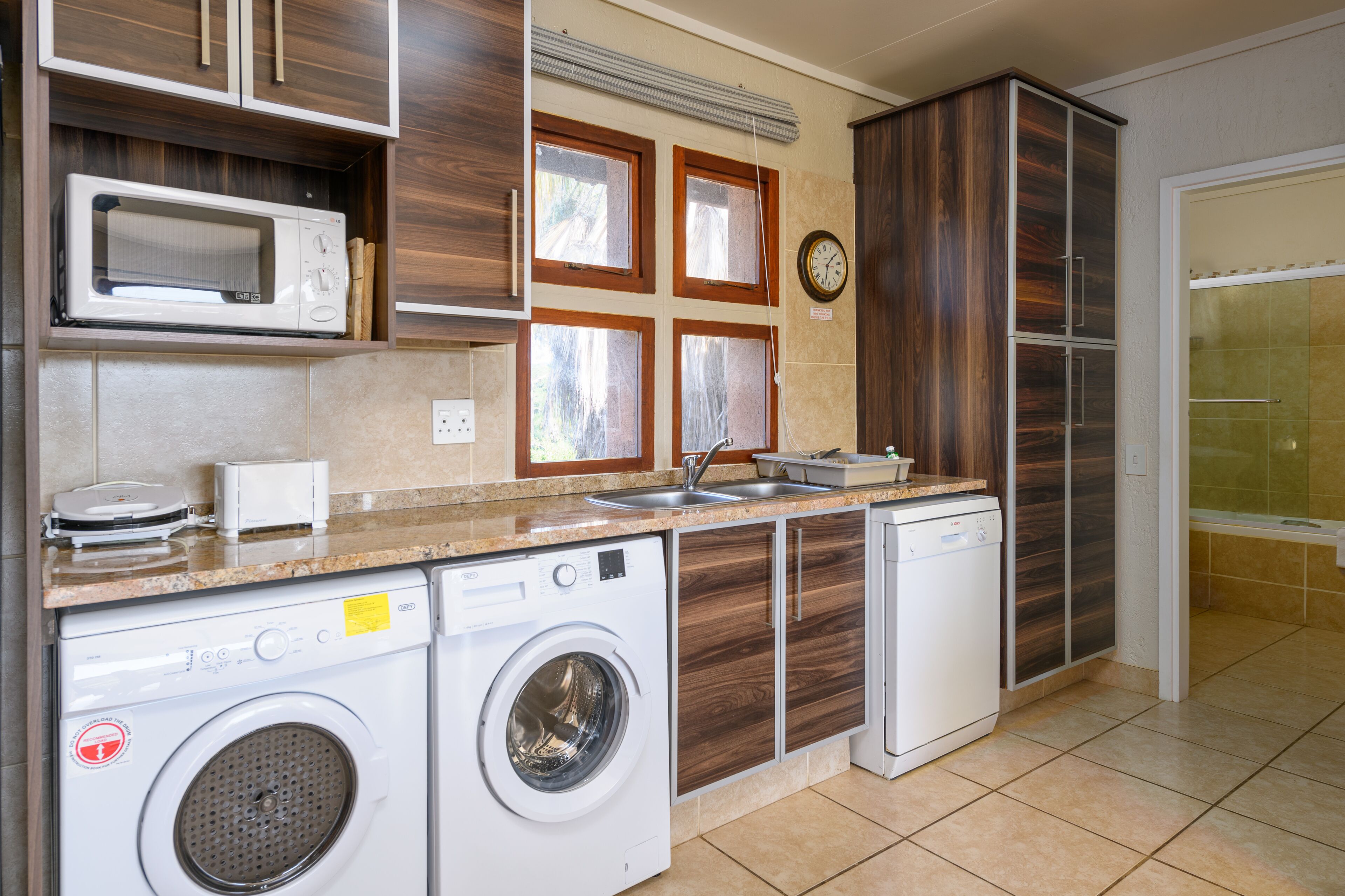 A compact kitchen with a microwave, washing machine, and dishwasher. There are wooden cabinets and a clock on the wall.