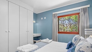 Ein Schlafzimmer mit Bett, Kleiderschrank, Fenster mit Vorhängen und Blick auf Bäume.