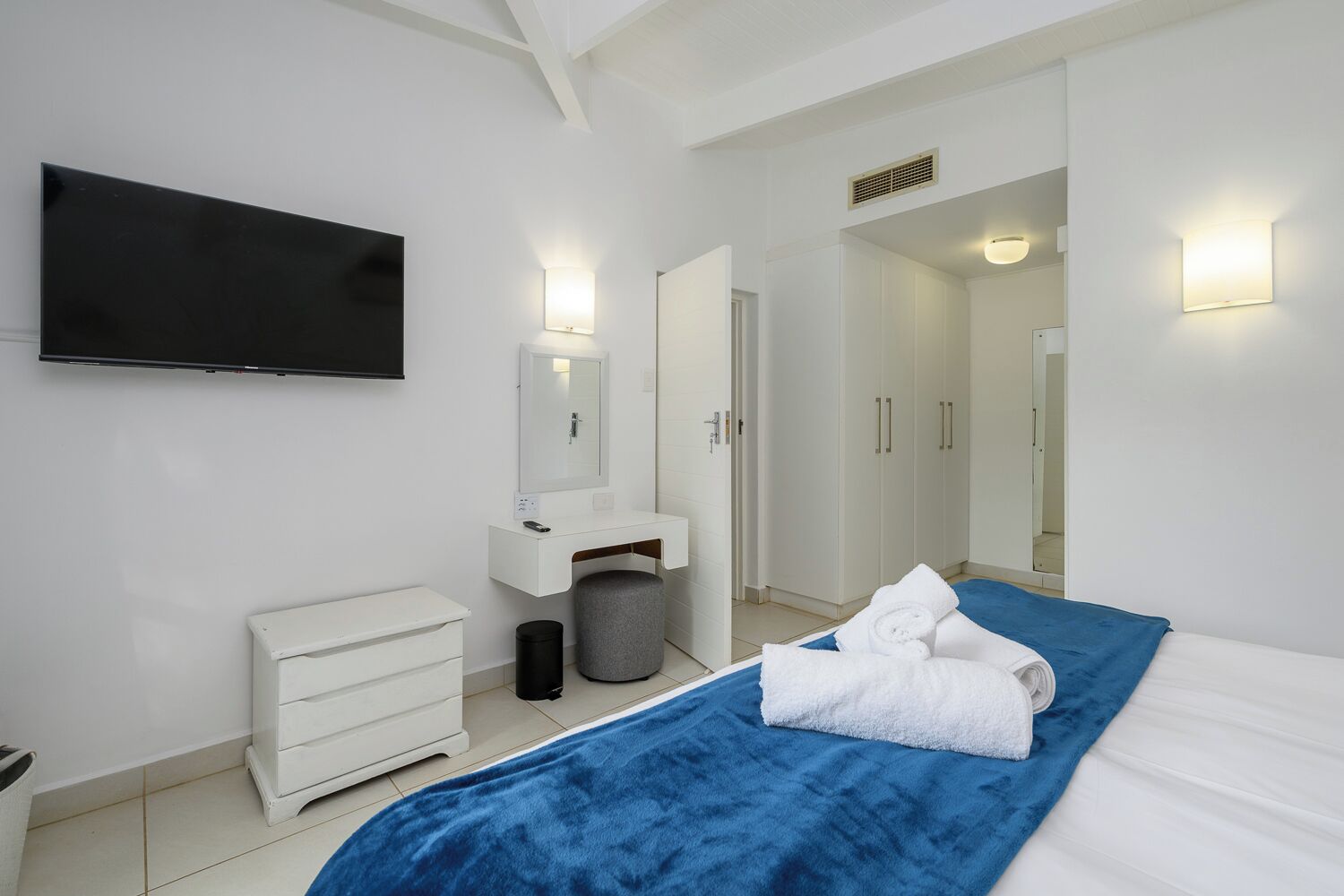 Habitació d'hotel moderna amb un llit, una petita taula de nit blanca, un tocador, un televisor de pantalla plana i un bany visible a través d'una porta oberta.