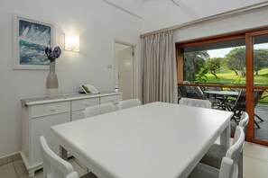 A room with a white table, chairs, a vase, a framed picture, a lamp, a mirror, and a view of a golf course through a glass door. - San Lameer Villa Rentals  2707 (Southbroom)