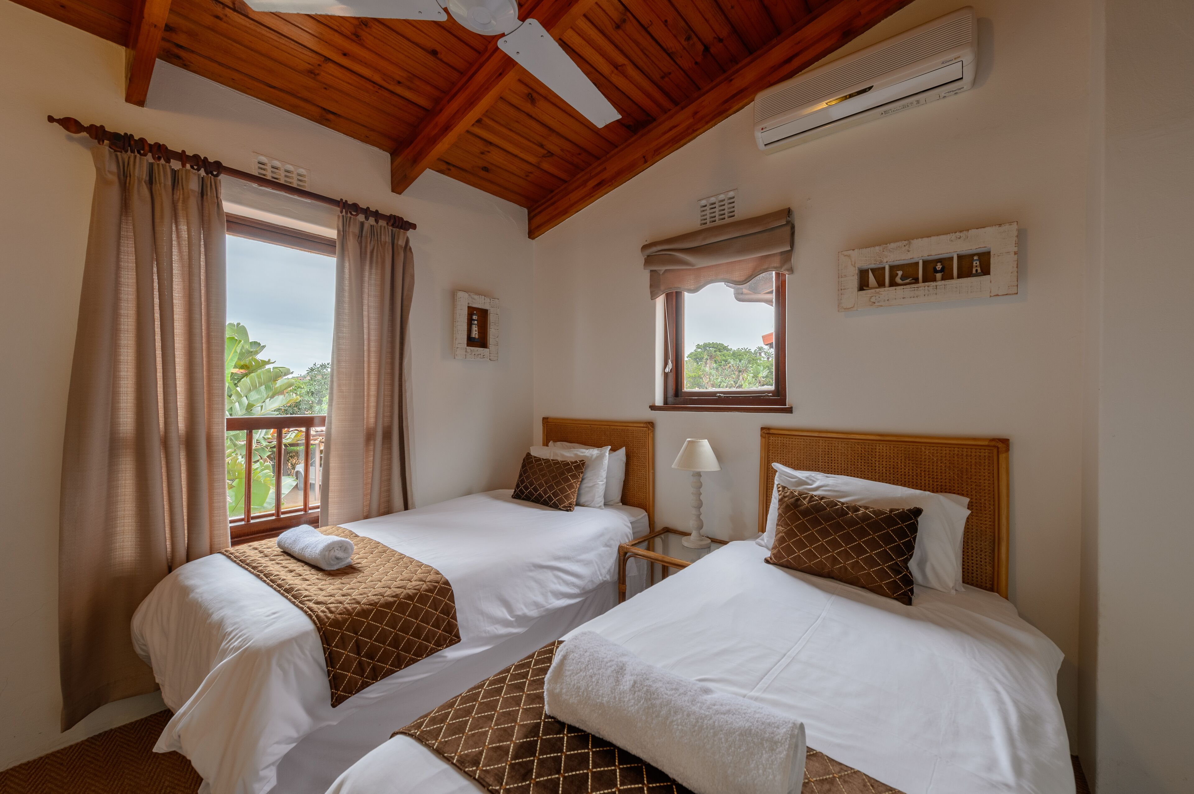 A hotel room with two beds, wooden ceiling beams, an air conditioning unit, a window with curtains, and a small table with a lamp.