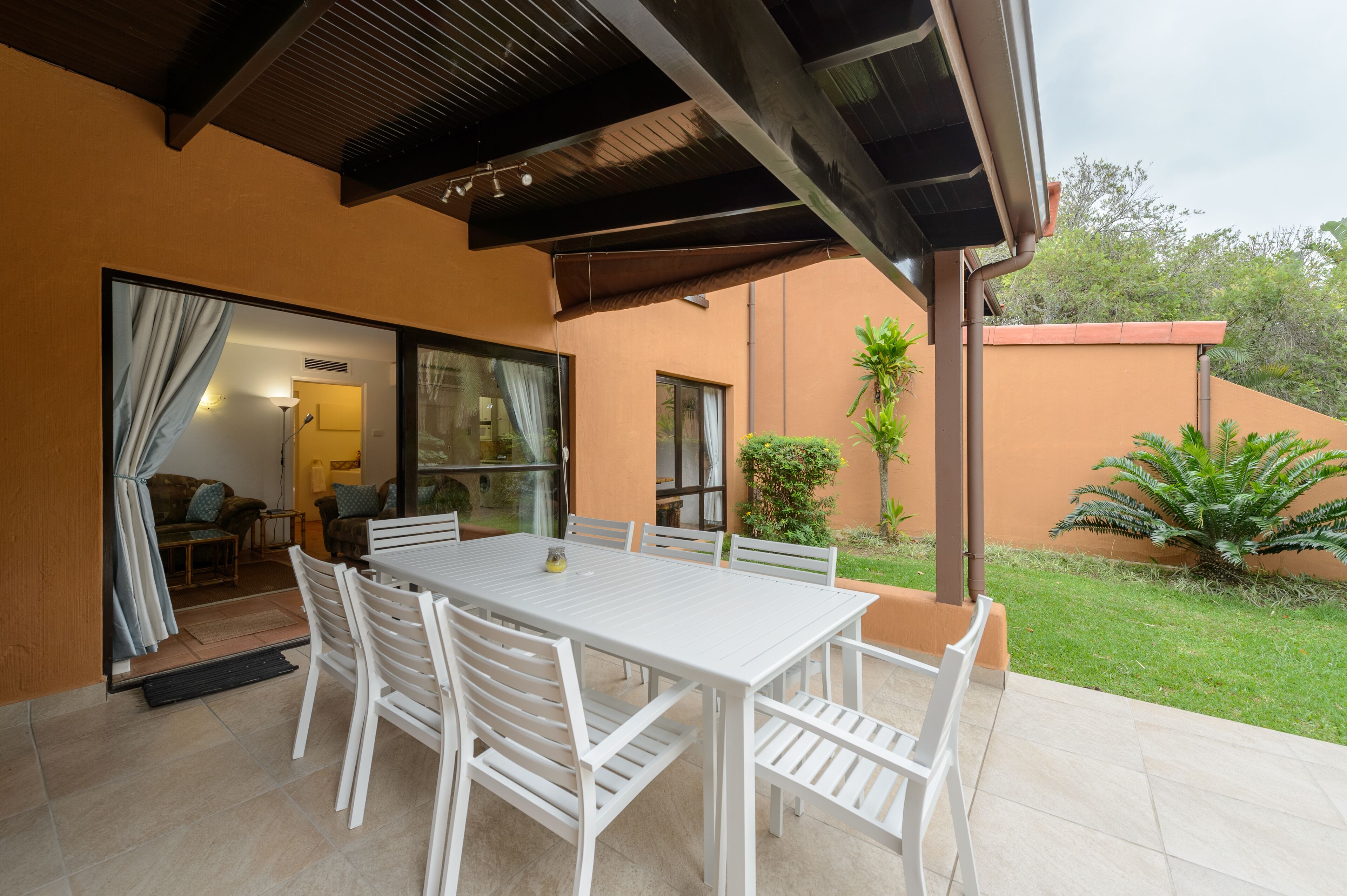 An outdoor patio with a white table and chairs, a covered area with a ceiling, and a view of a garden with plants and a tree.