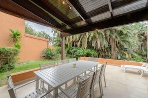 Un patio al aire libre con mesa y sillas, rodeado de abundante vegetación y palmeras.
