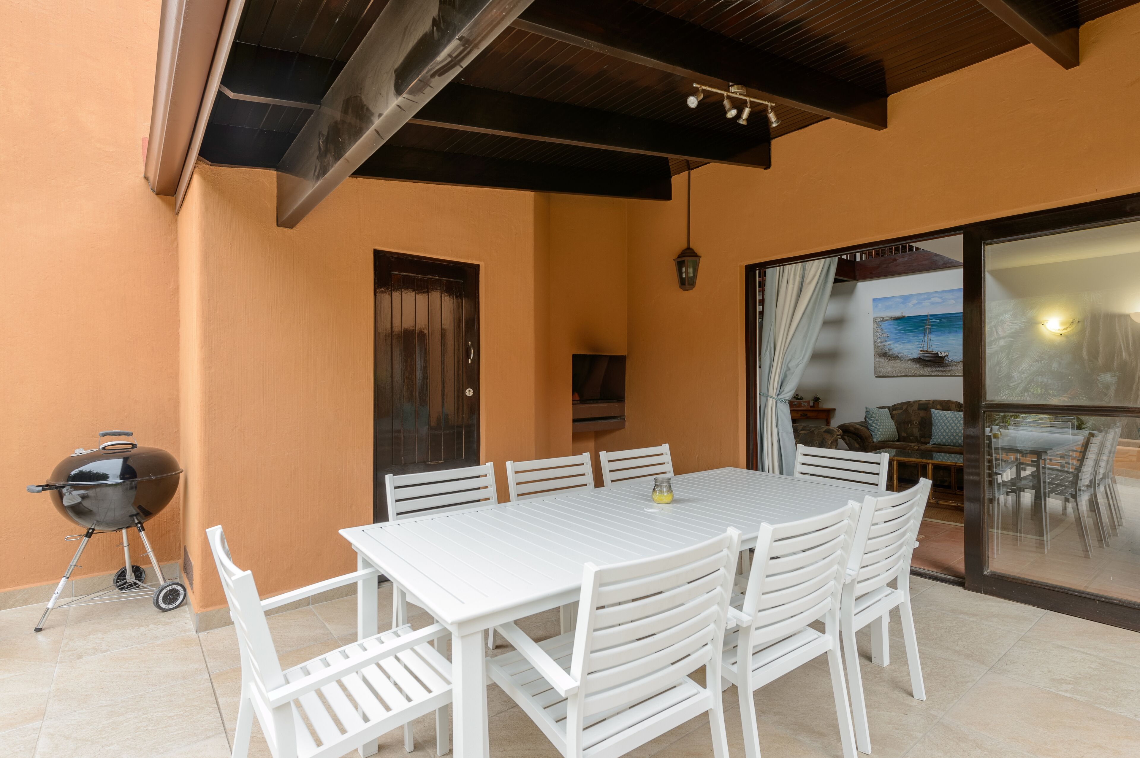 An outdoor patio with a white table and chairs, a barbecue grill, and a sliding glass door leading to an indoor area with a painting and furniture.