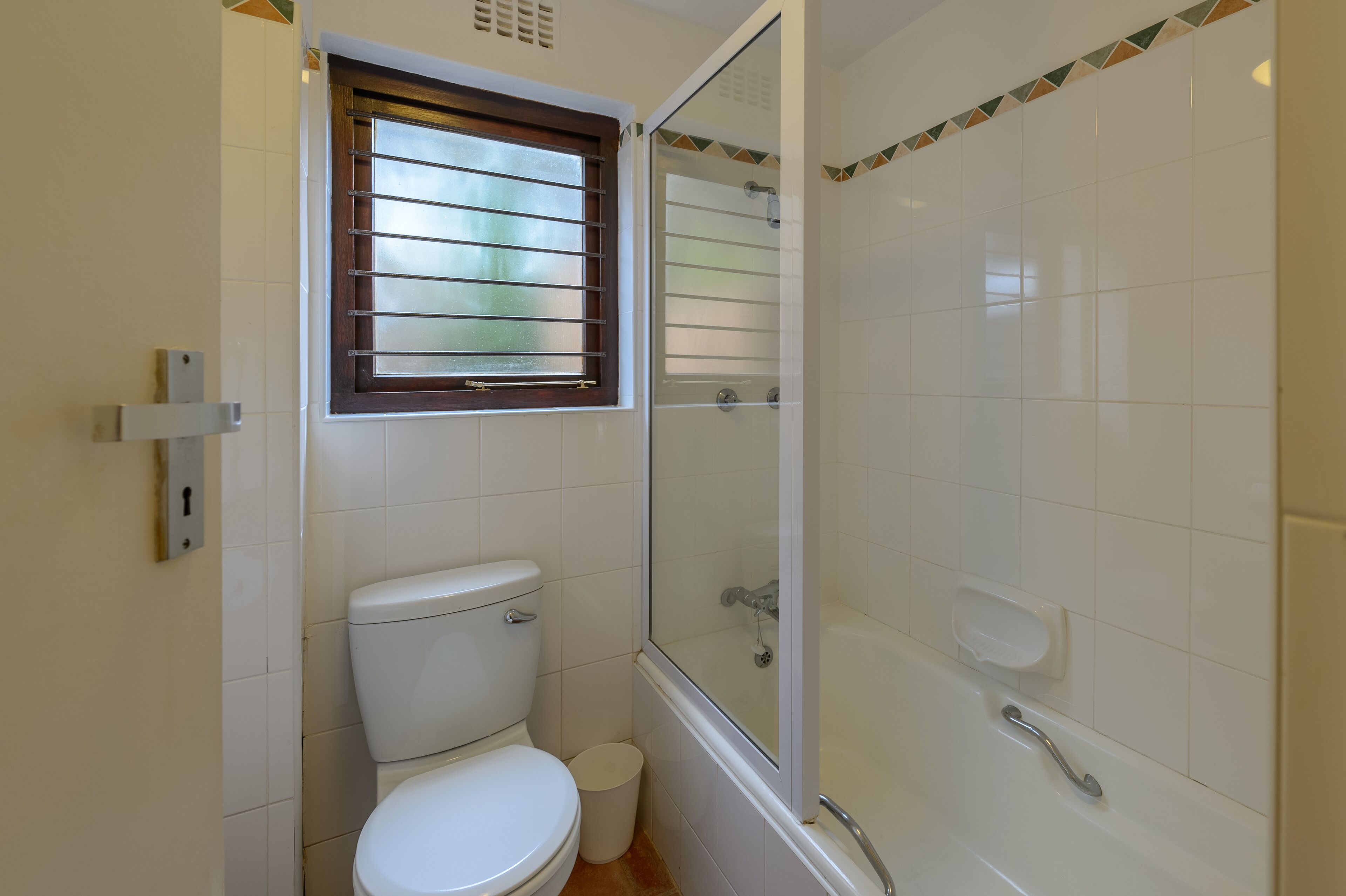 A bathroom with a toilet, a window with blinds, a shower with a glass door, and a white sink.