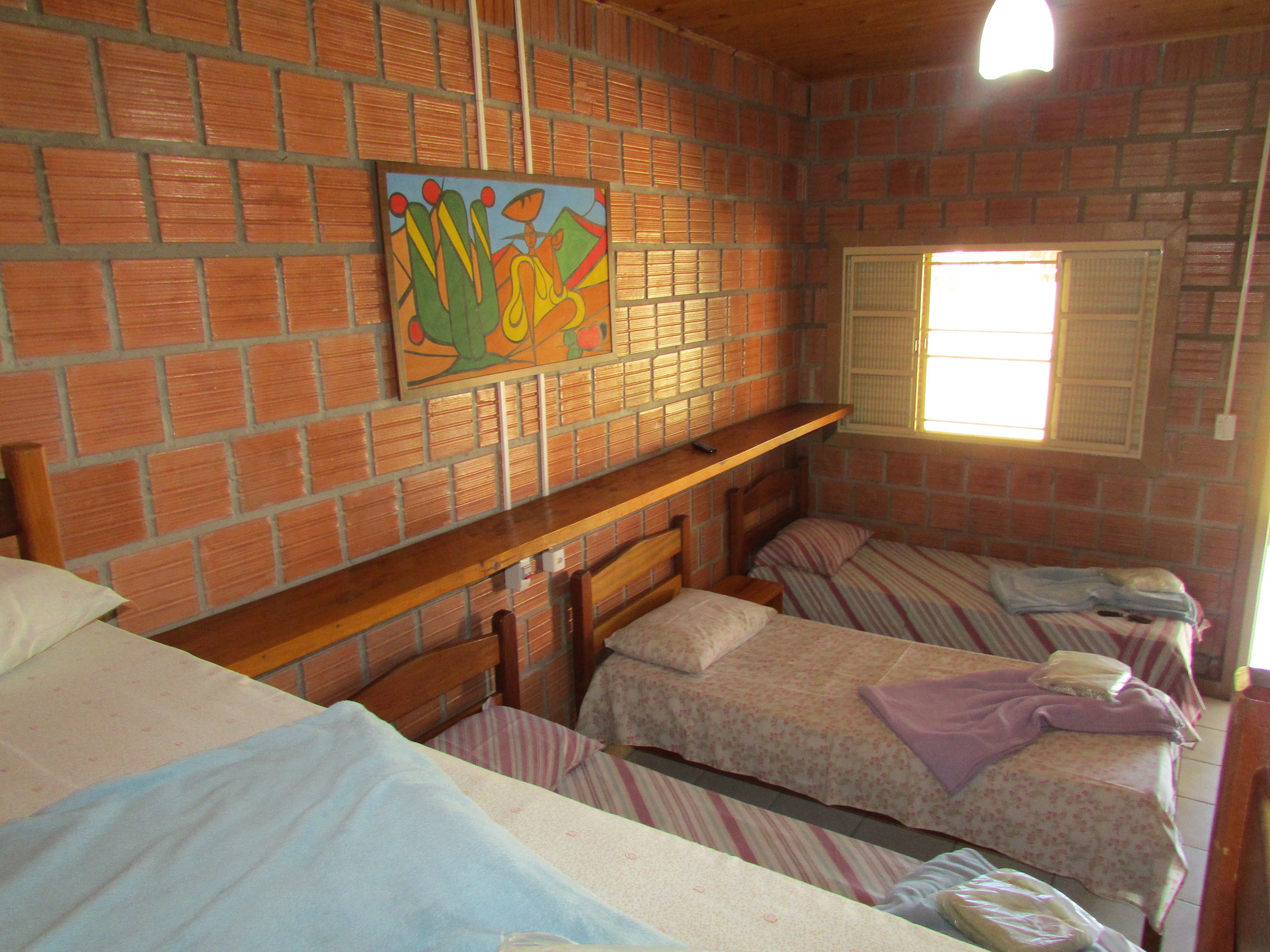 A room with brick walls, a wooden ceiling, and a window with blinds. There are three beds with striped bedding and a painting on the wall.