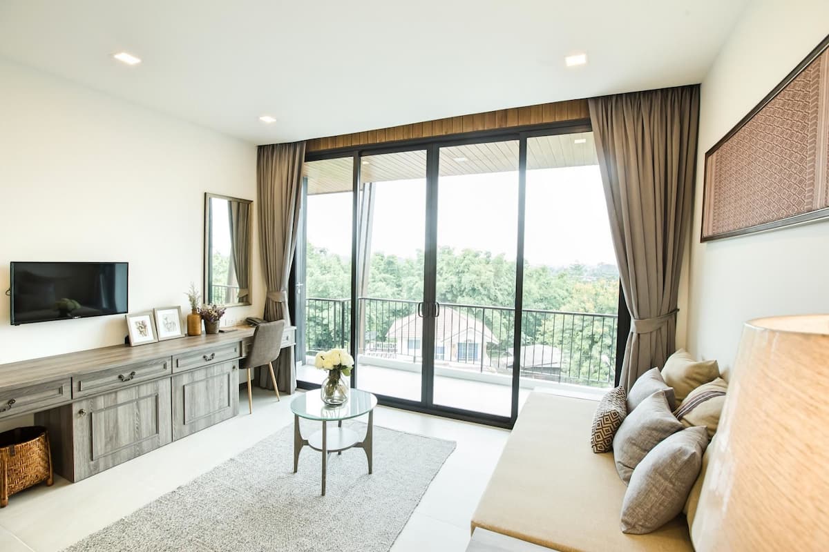 Uma sala de estar moderna com sofá, televisão, área de jantar com mesa e cadeiras, e varanda com vista para o verde.