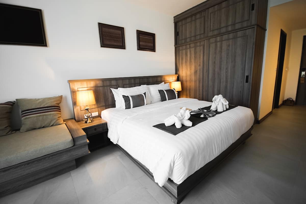 Quarto de hotel moderno com uma cama grande, um sofá, uma mesa de cabeceira com abajur e parede de painéis de madeira.