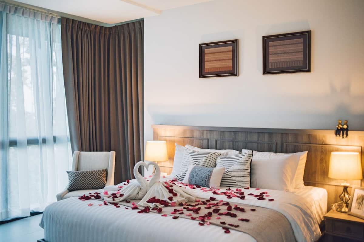 Quarto de hotel com uma cama grande, duas luminárias de cabeceira, um espaldar e uma janela com cortinas.