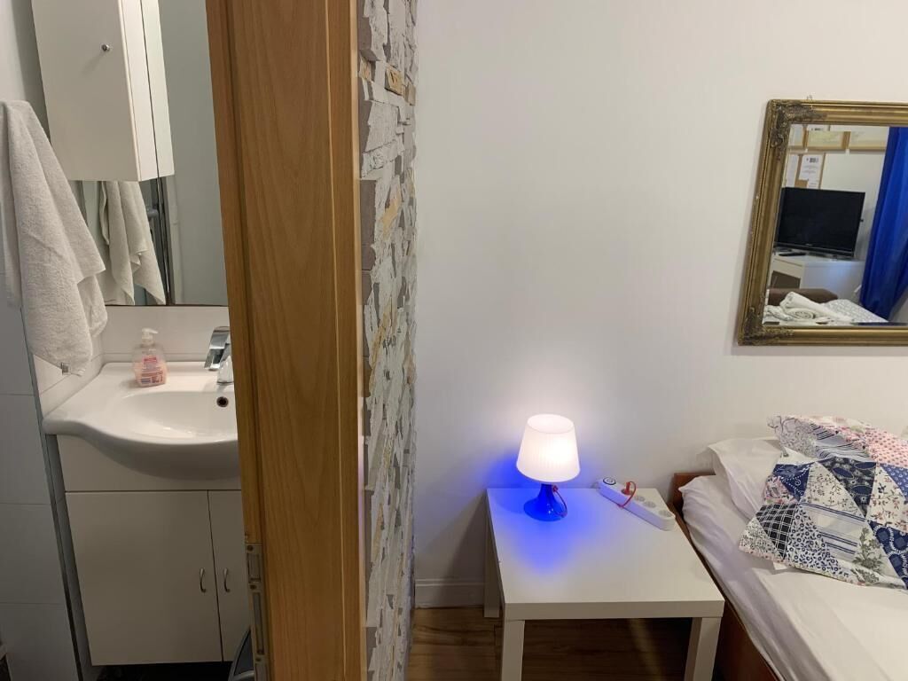 A bedroom with a bedside table, a lamp, a bed with a quilt, a mirror, and a bathroom with a sink and towel.