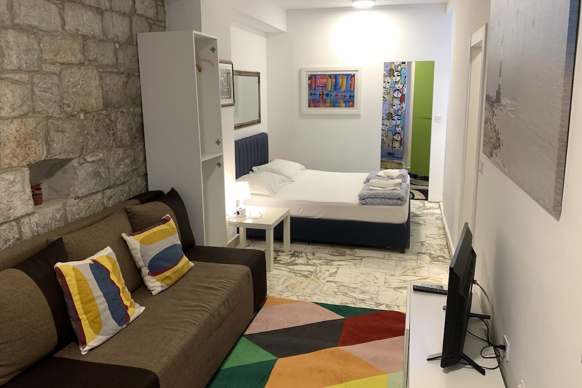 A compact hotel room with a stone wall, a bed with a blue headboard, a white bedside table with a lamp, a colorful rug, and a television.