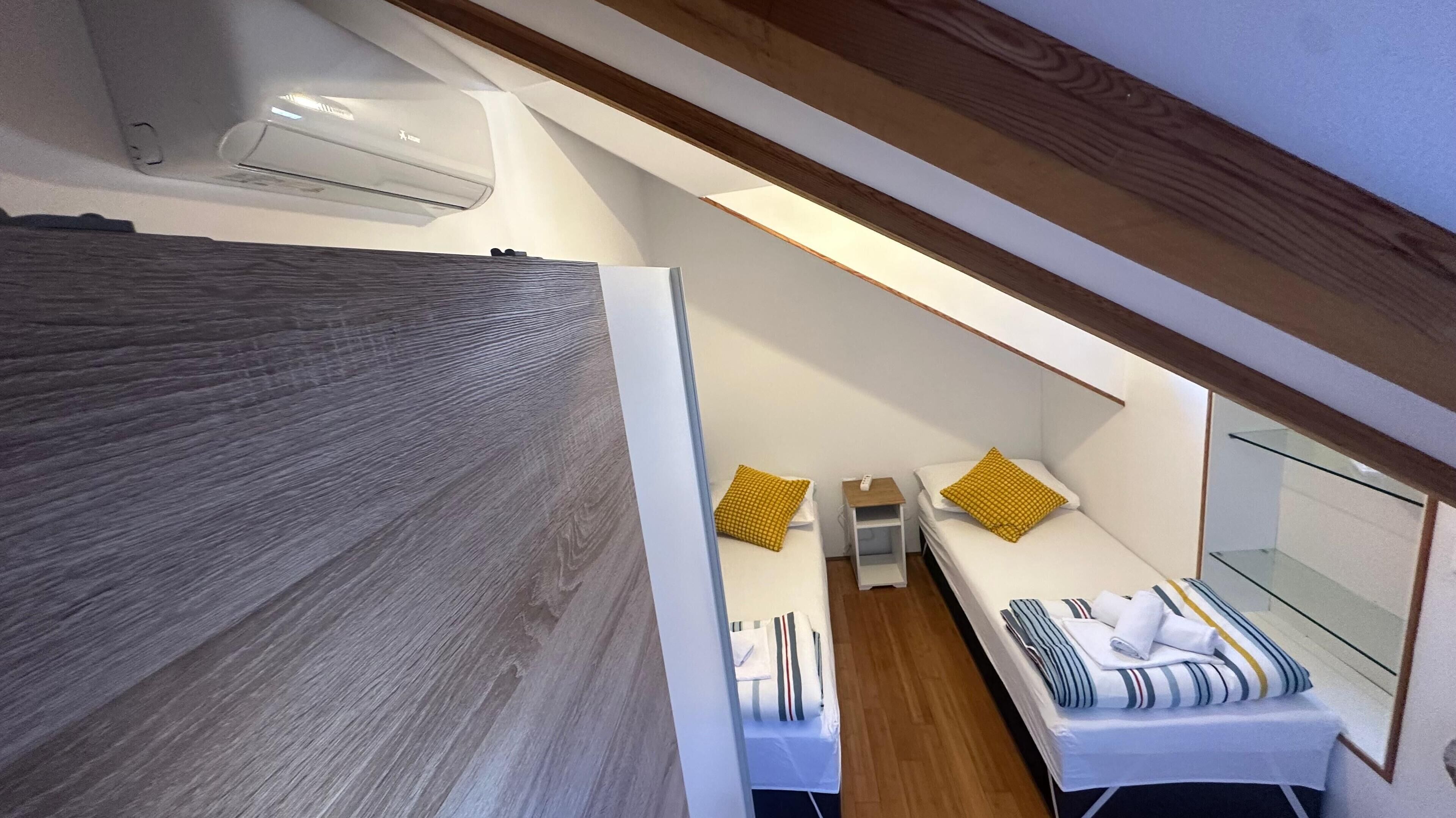 Ein gemütliches Schlafzimmer mit schrägem Dach, Holzboden, zwei Betten mit gestreifter Bettwäsche und einem kleinen Nachttisch mit Lampe.