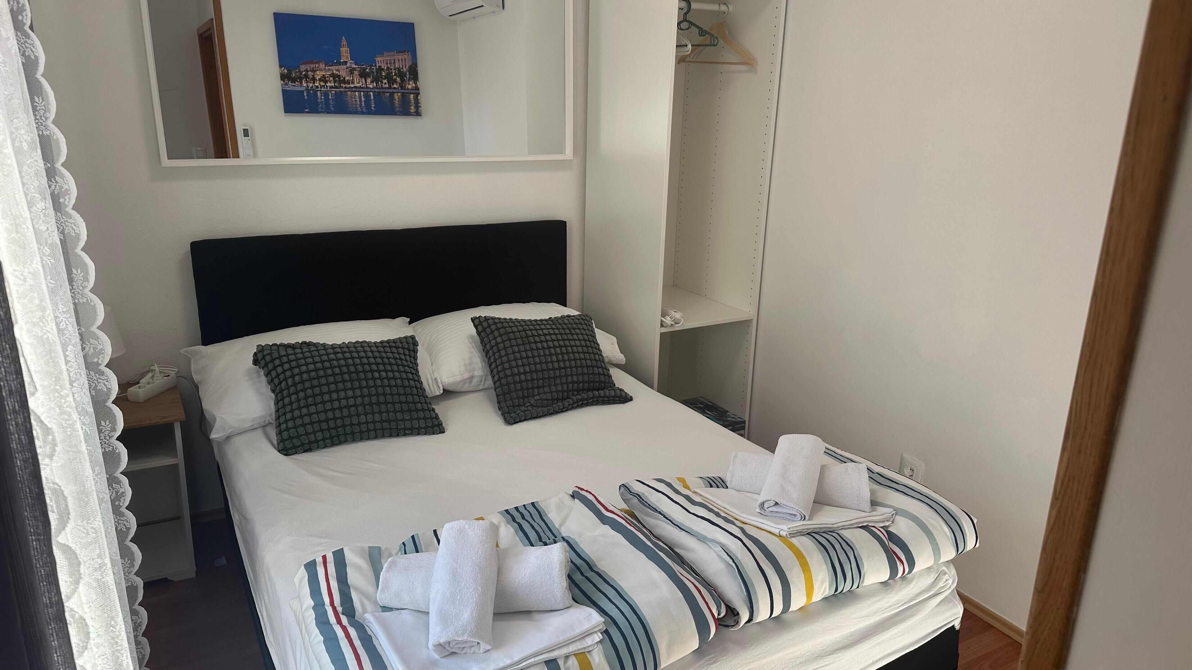 A neatly made bed with striped bedding and two pillows, a bedside table with a phone, a wall-mounted painting, and a hanging coat rack.