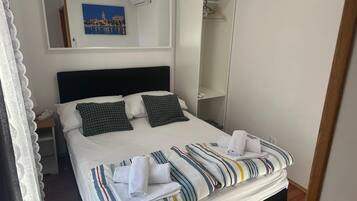 A neatly made bed with striped bedding and two pillows, a bedside table with a phone, a wall-mounted painting, and a hanging coat rack.
