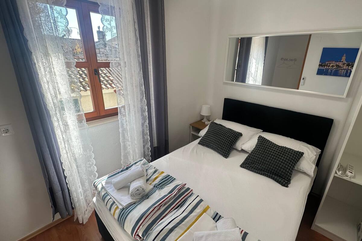 A neatly made bed with striped bedding and two pillows, a bedside table with a lamp, a window with curtains, and a wall-mounted mirror.