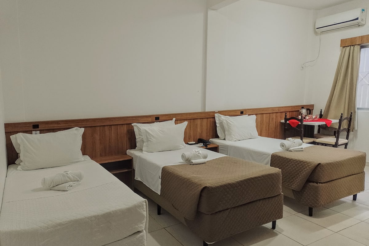 Habitación de hotel con dos camas individuales, una cama doble, una mesita con mantel rojo y un equipo de aire acondicionado en la pared.