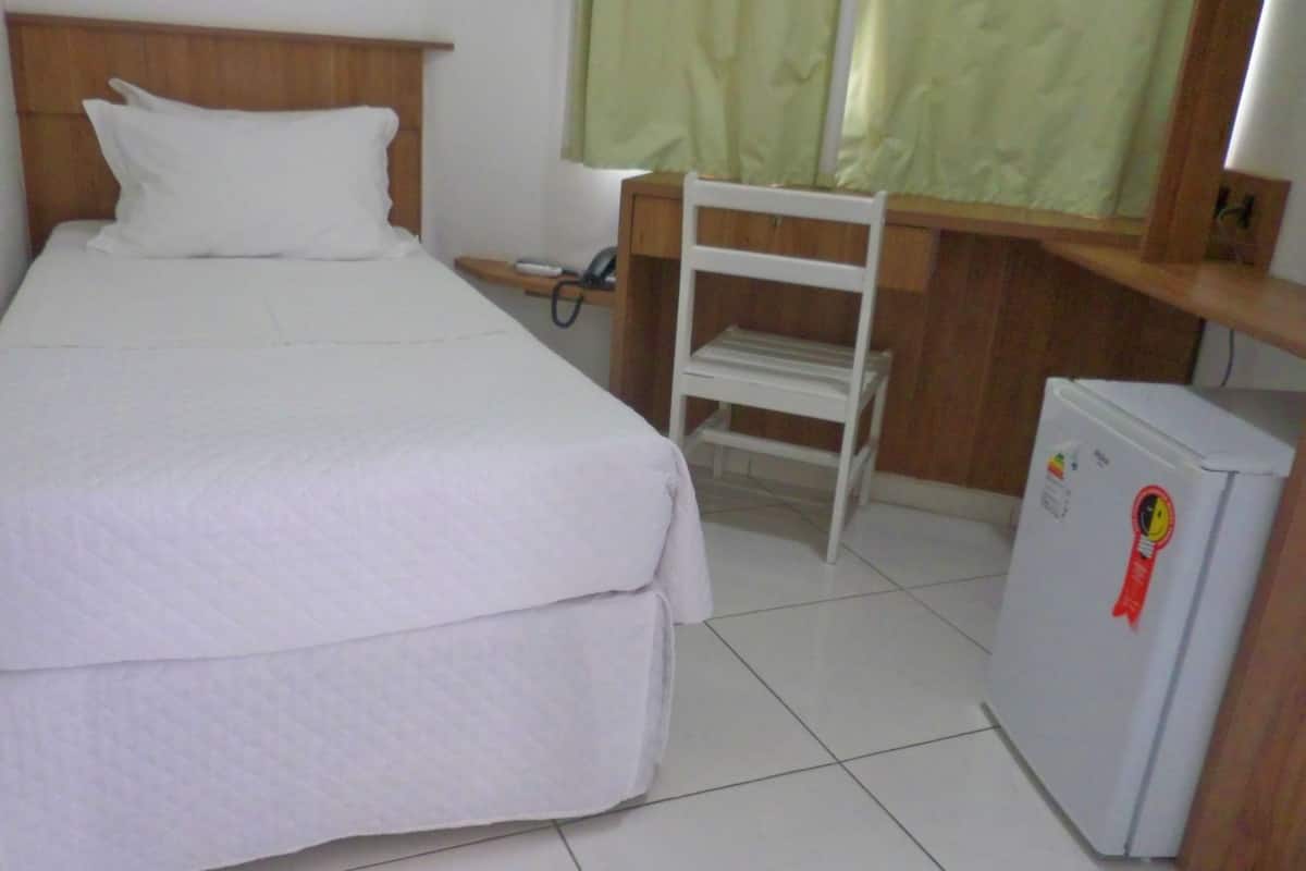 Habitación individual con una cama de madera, cabecera de madera, mesita de noche con lámpara, refrigerador blanco pequeño y una ventana con cortinas verdes.
