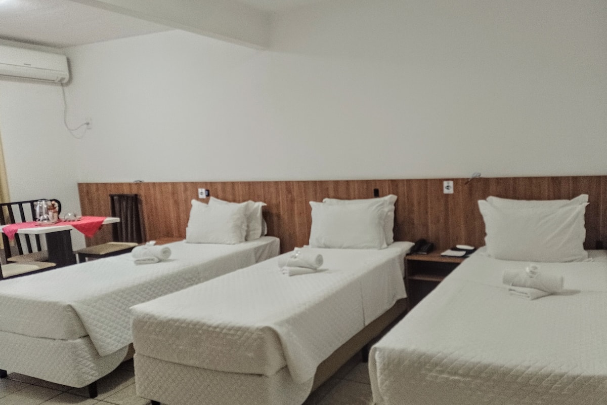 Una habitación de hotel con tres camas individuales, un cabecero de madera, una mesita con mantel rojo y una silla.