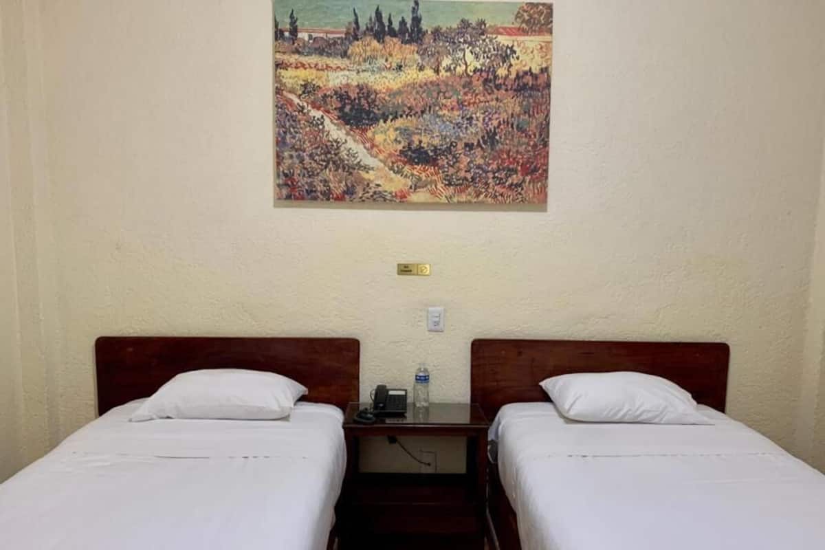 Habitación de hotel con dos camas individuales, una mesita de noche con un teléfono y una botella, y un cuadro en la pared.