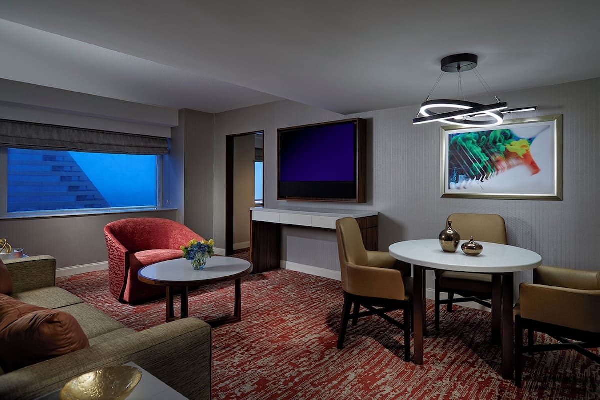 A modern hotel room with a flat-screen TV, a painting, a red chair, a round table, and a red carpet.