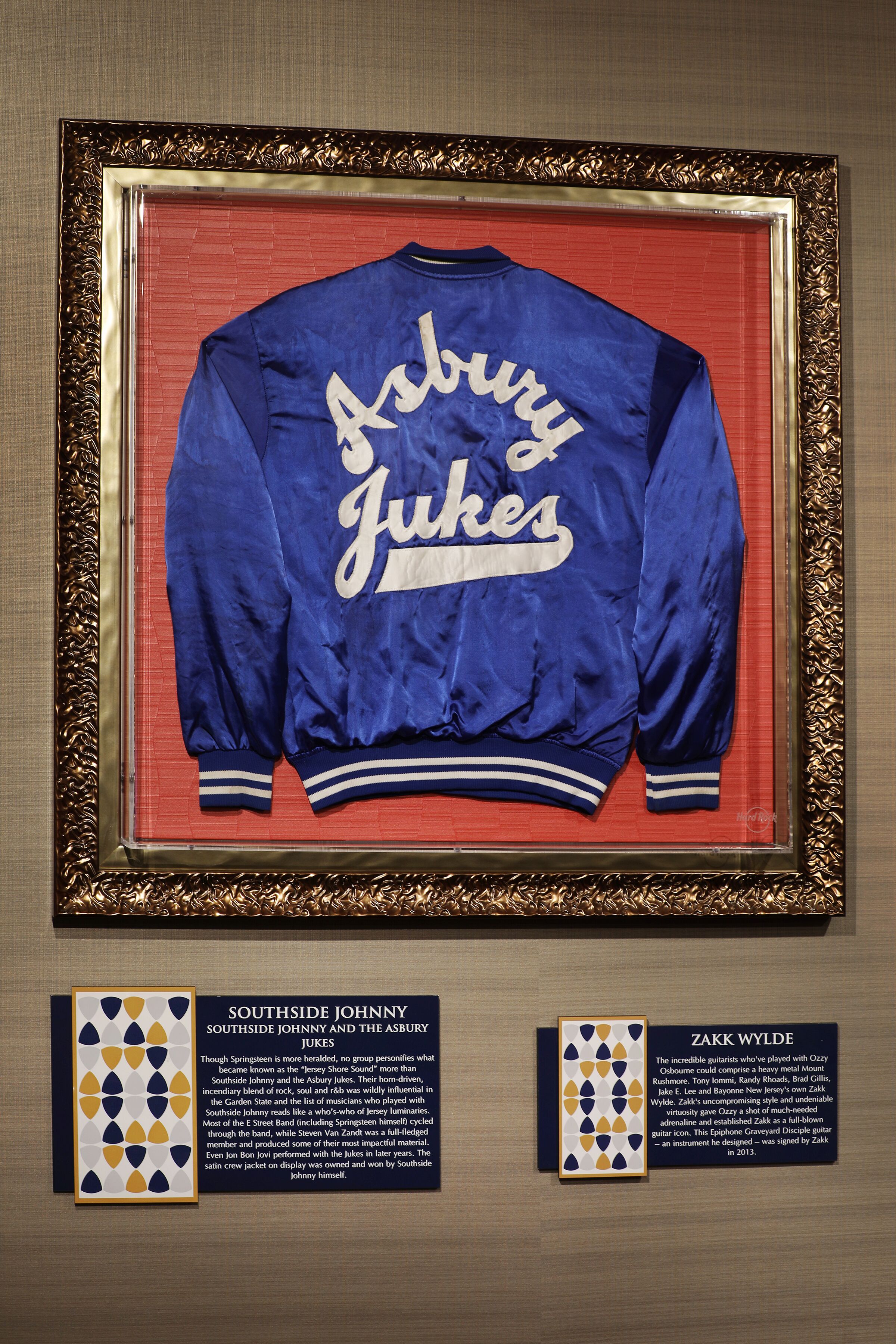 Una chaqueta universitaria Auburn & Jukes enmarcada con un marco dorado, exhibida en una pared con dos placas informativas debajo.