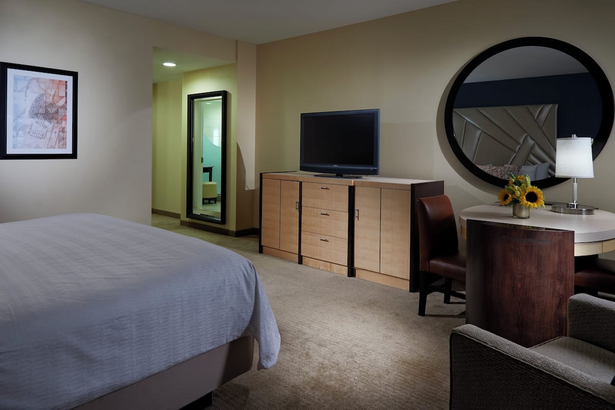 A hotel room with a bed, a TV on a wooden cabinet, a round mirror, a desk with a lamp, and a chair.
