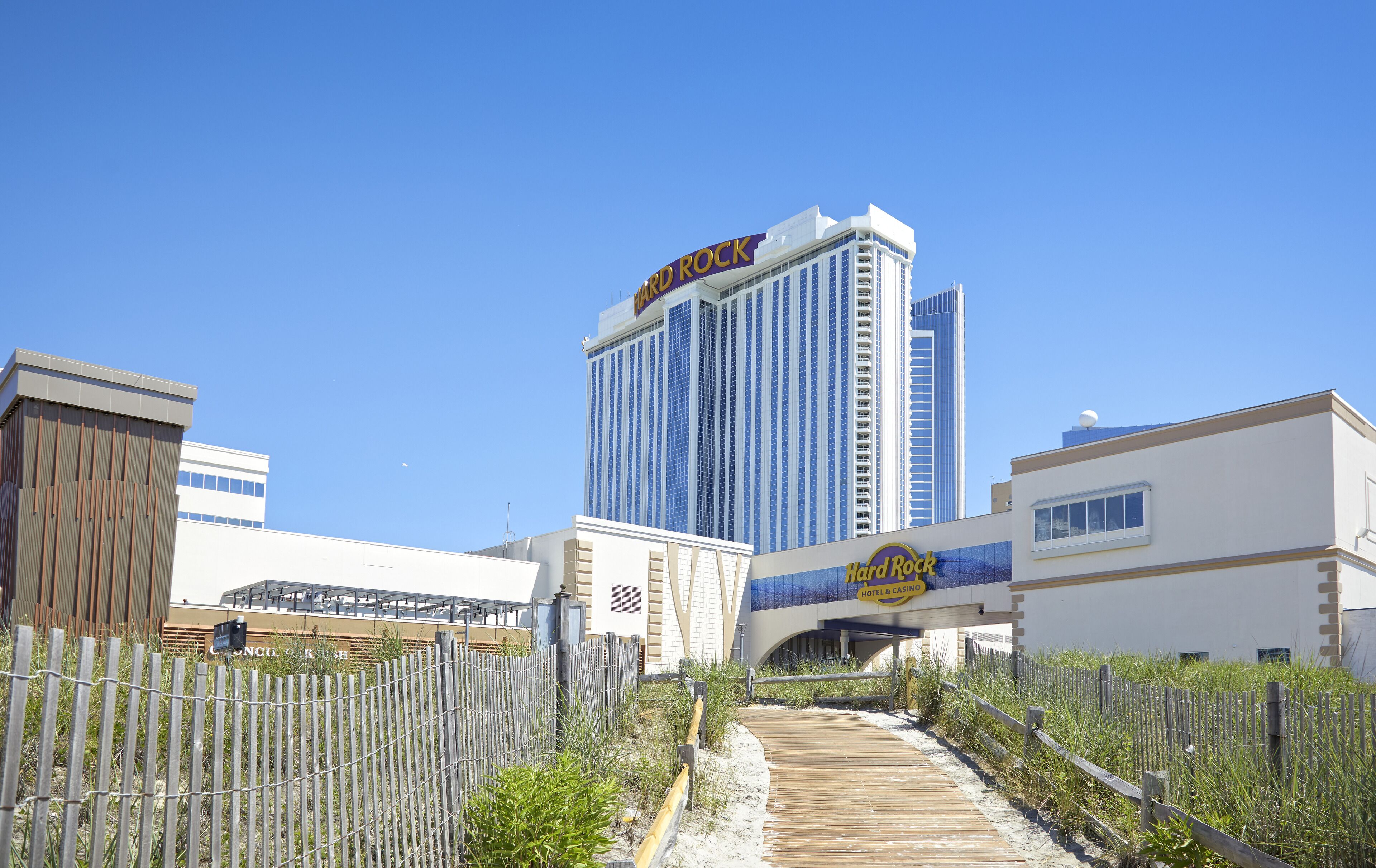 A hotel with a prominent 'Hard Rock' sign, a wooden boardwalk leading to the entrance, and a clear blue sky.