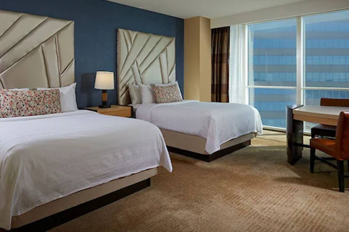 A hotel room with two beds, a desk, and a large window offering a city view.