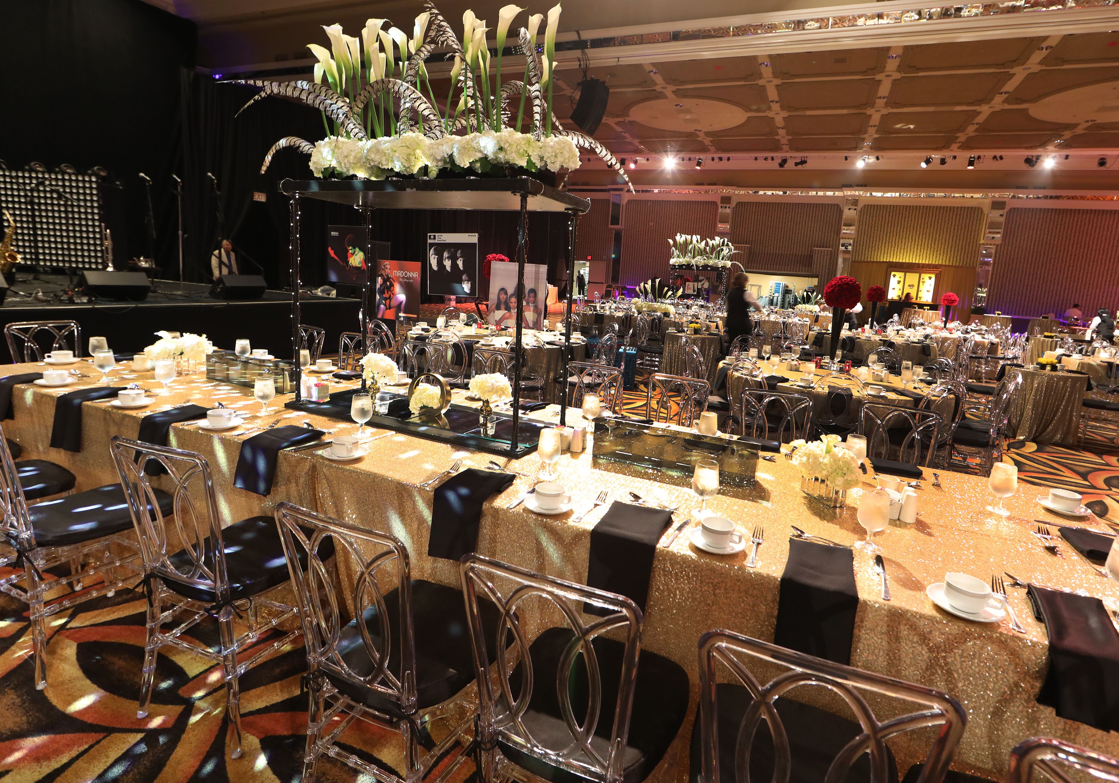 A banquet hall with long tables set for a formal event, featuring clear chairs, gold tablecloths, and floral centerpieces.