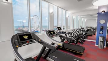 Un gimnasio con cintas de correr y equipo de ejercicio, amplias ventanas con vista a un muelle y una noria, y un reloj en la pared.