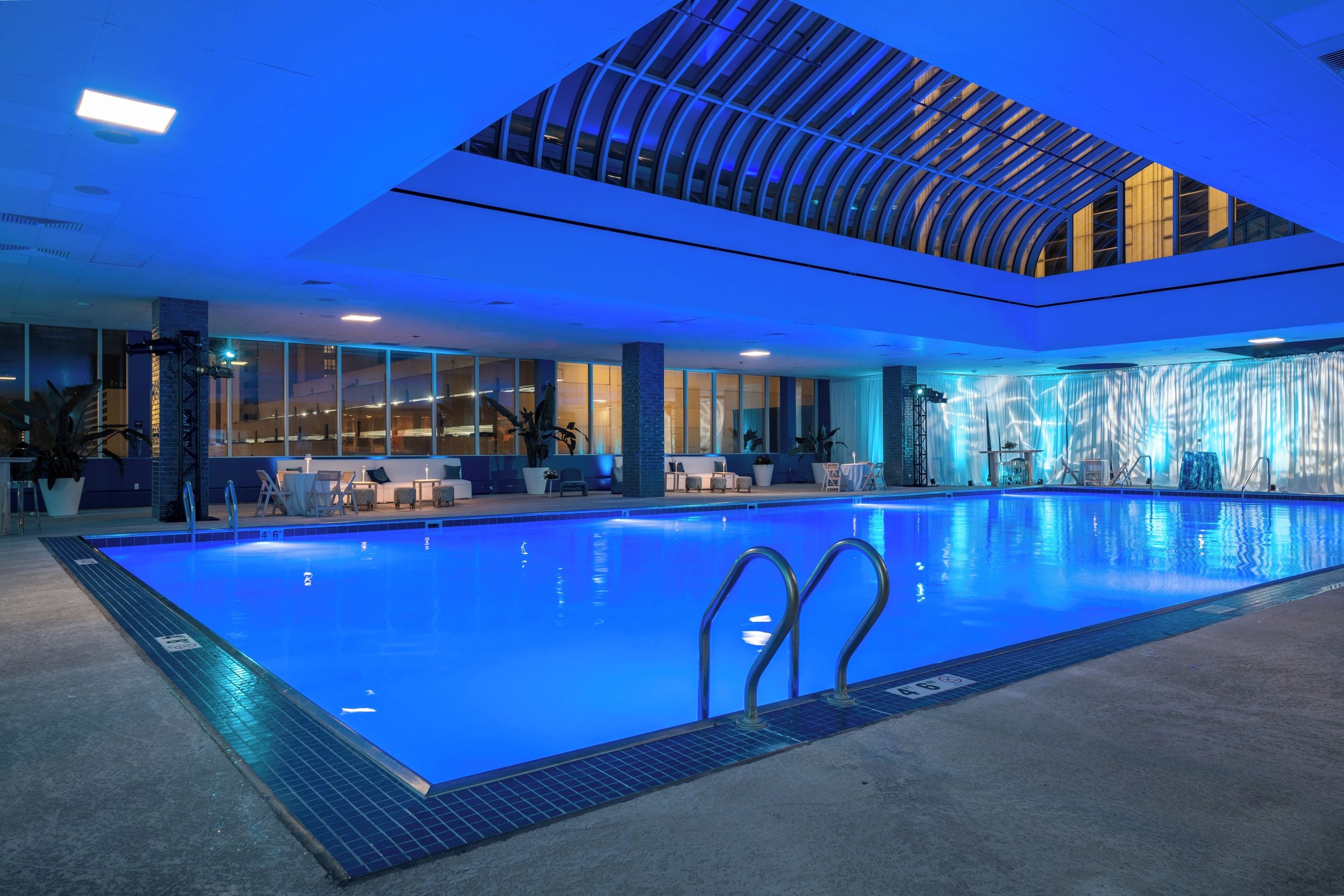 A large indoor swimming pool with clear blue water, surrounded by lounge areas with seating and potted plants.