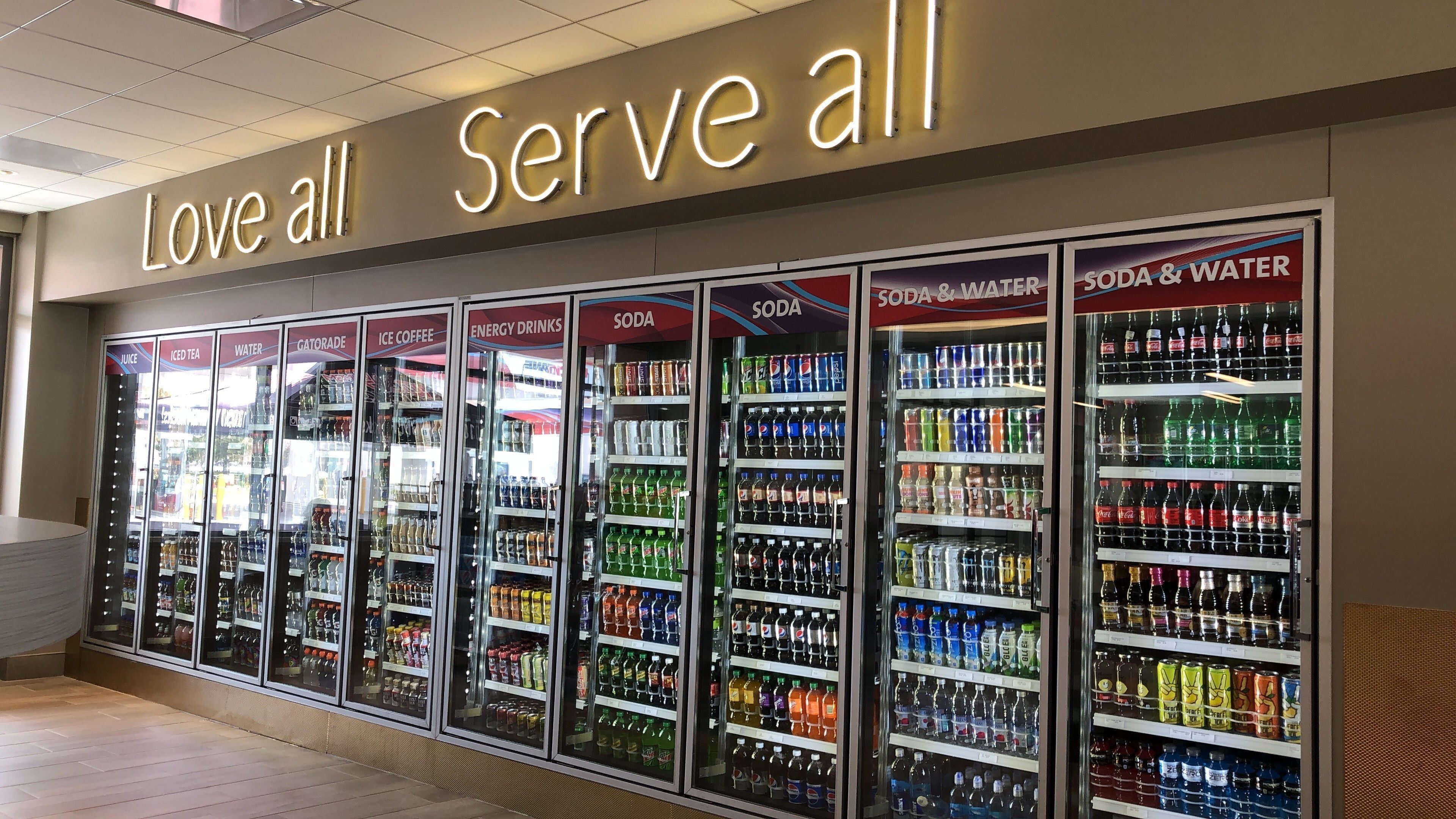 A row of refrigerated beverage dispensers with various drink options, including sodas, juices, and energy drinks, under a sign that reads "Love all Serve all".