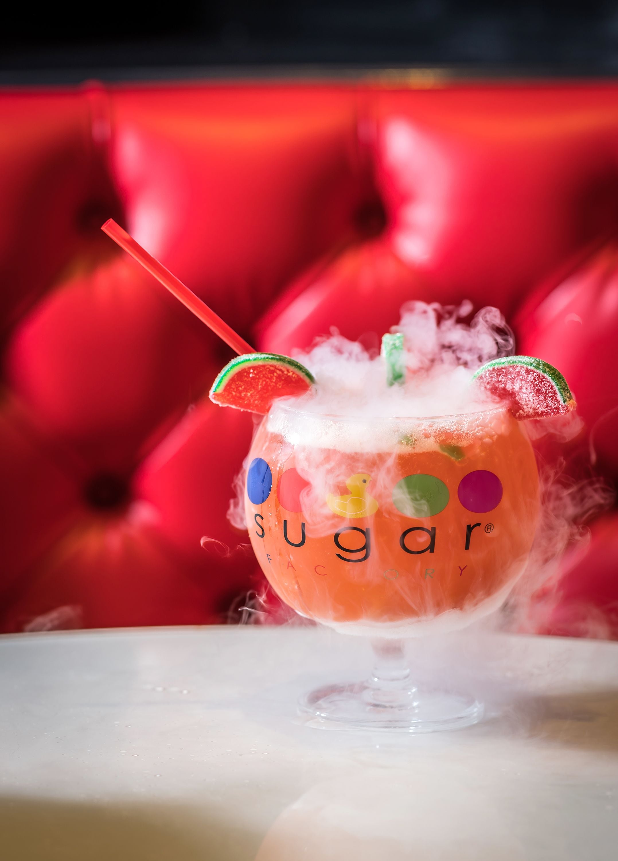 A cocktail with a straw and lime slice garnish, labeled "sugar" on the glass, with a red upholstered background.