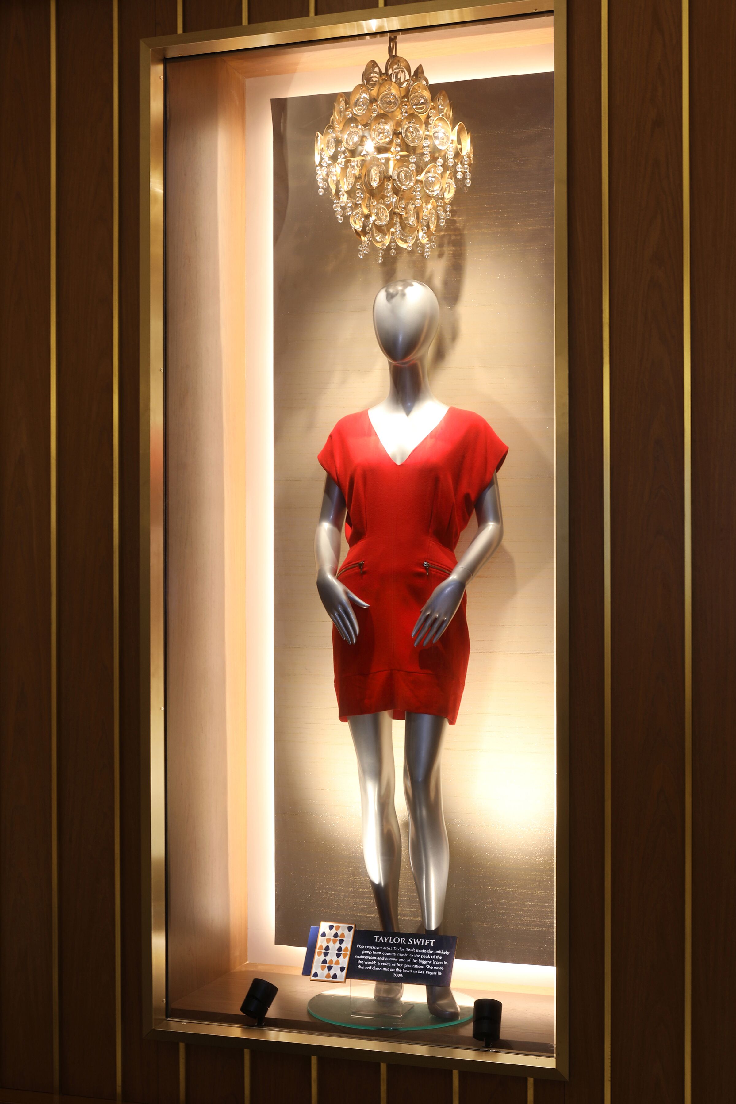 A mannequin in a red dress inside a framed display with a chandelier above.
