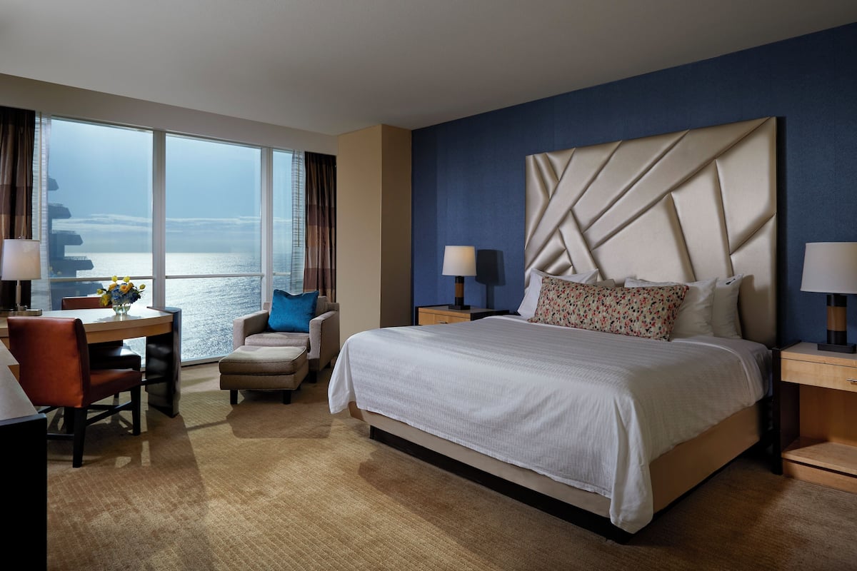 A hotel room with a large bed, a desk with a chair, a view of the sea, and a nightstand with a lamp.