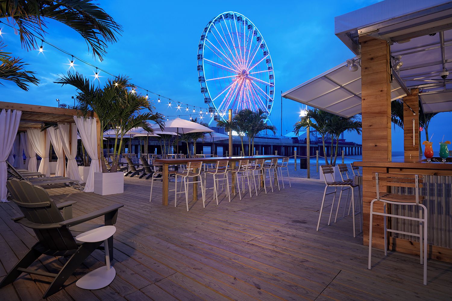 An outdoor bar area with wooden tables and chairs, a Ferris wheel in the background, and palm trees.