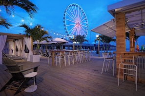 An outdoor bar area with wooden tables and chairs, a Ferris wheel in the background, and palm trees. - Hard Rock Hotel & Casino Atlantic City (Atlantic City)