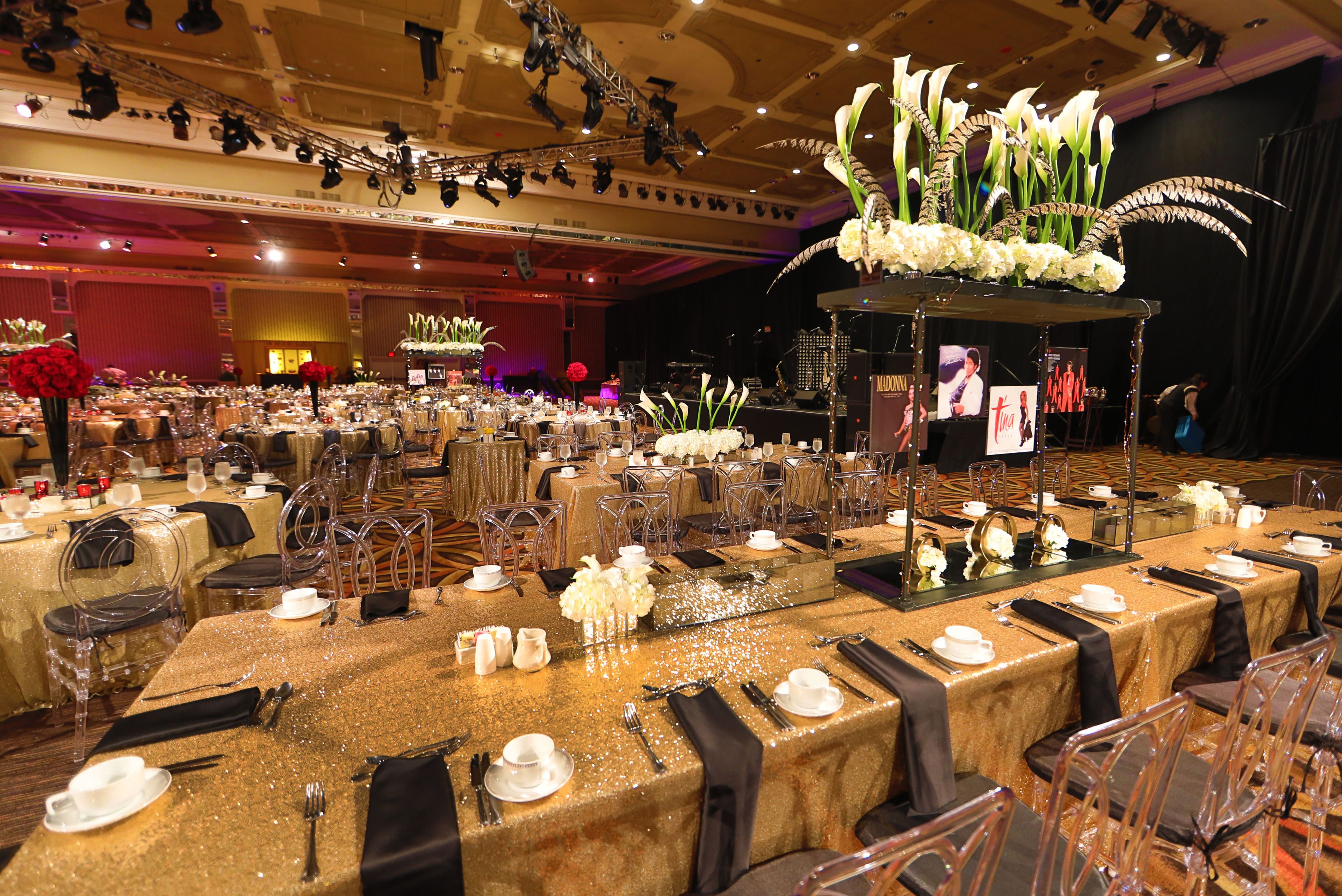 A banquet hall with tables set for a formal event, featuring clear chairs, gold tablecloths, and floral centerpieces.