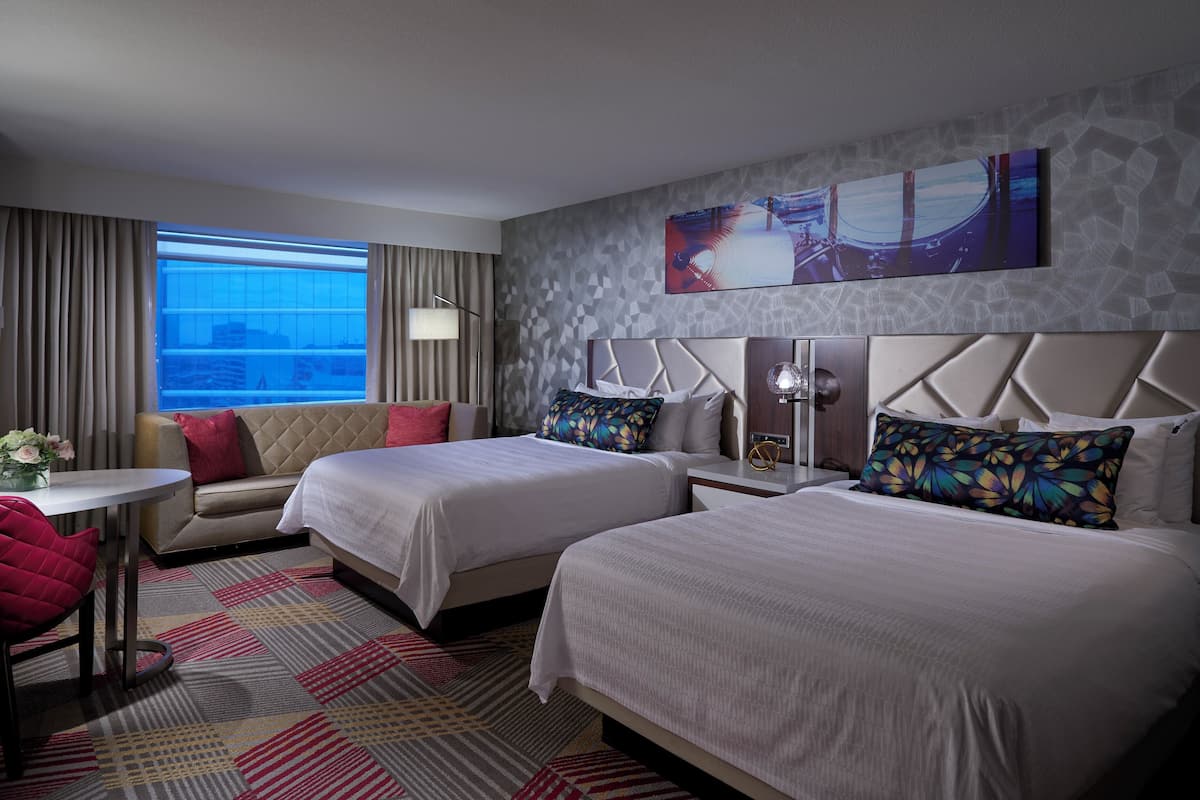 A hotel room with two beds, a sofa, a small table, and a chair. There is a large window with curtains, a wall-mounted artwork, and a patterned carpet.