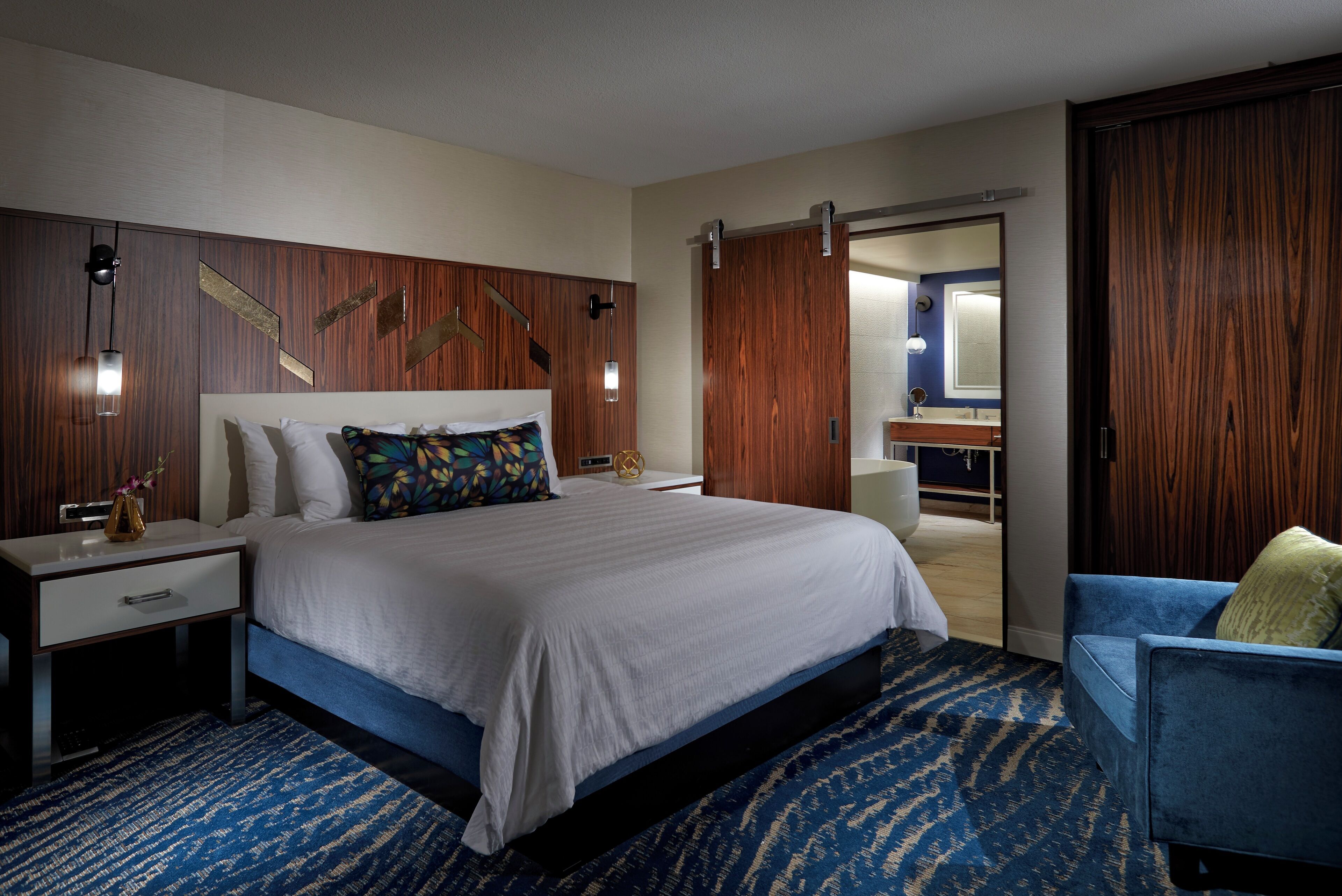 Camera d'albergo moderna con un letto grande, un comodino, una scrivania, una poltrona blu e un'armadio in legno.