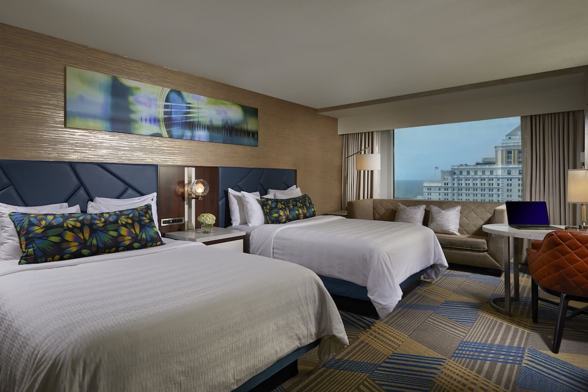 A hotel room with two beds, a sofa, a laptop on a side table, and a view of a cityscape.